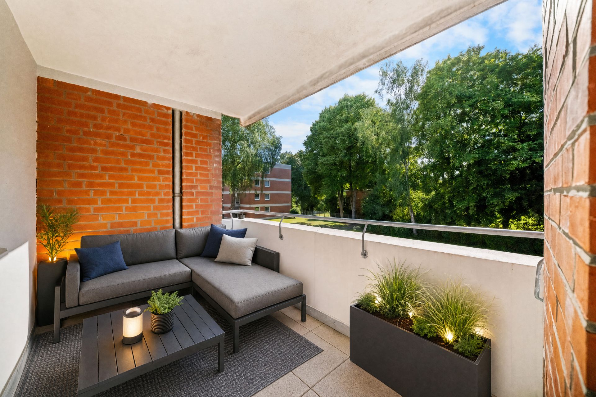 Überdachter Balkon mit grauem Sofa, Couchtisch, Pflanze und Blick auf Bäume.