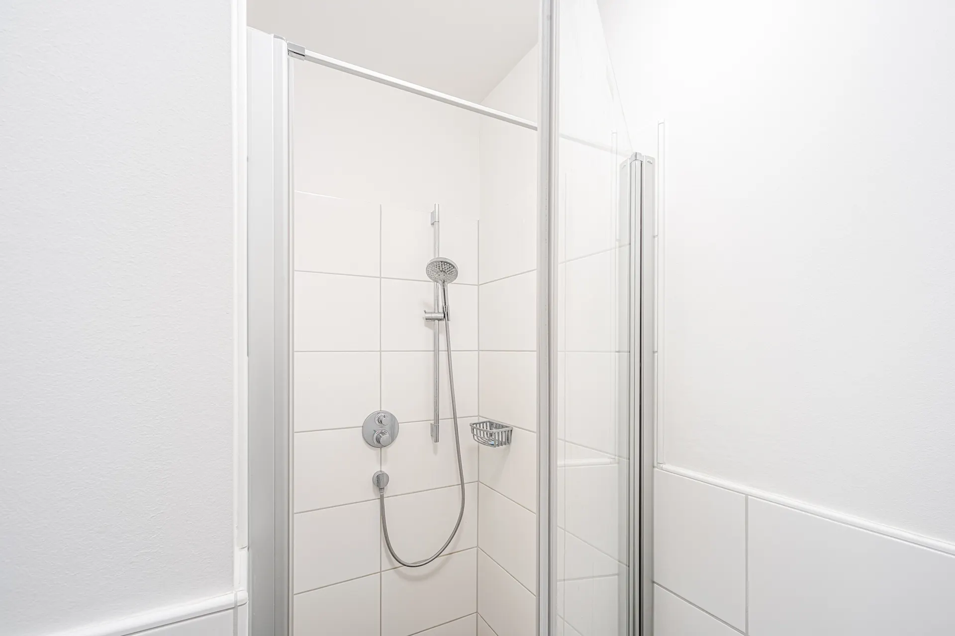 Weiße, geflieste Dusche mit Glastür und verstellbarem Duschkopf an der Wand.