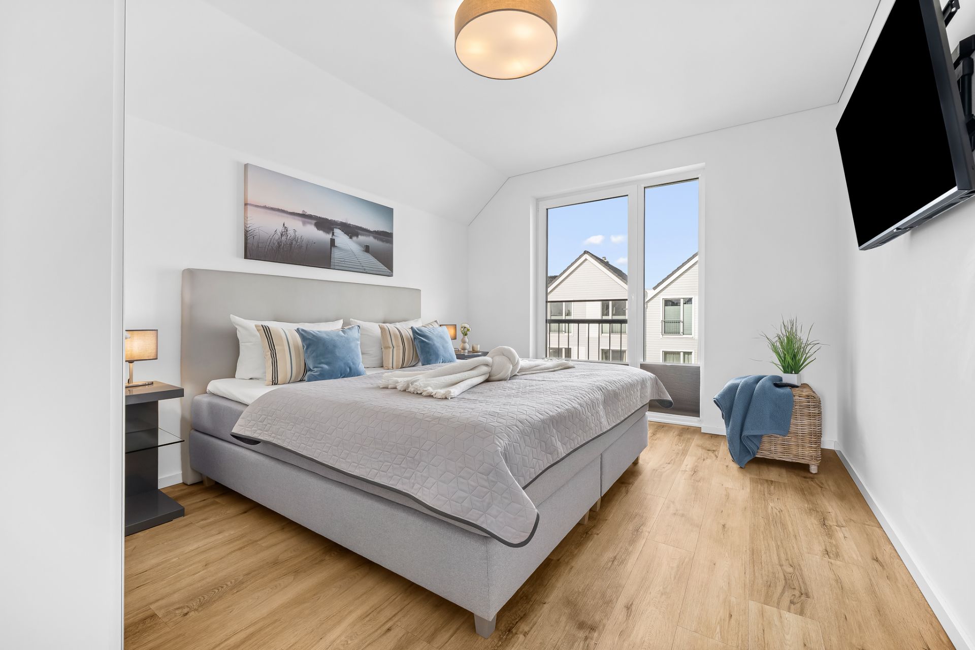 Helles Schlafzimmer mit großem Bett, Nachttischen, Wandbild, Fernseher und Balkonzugang.