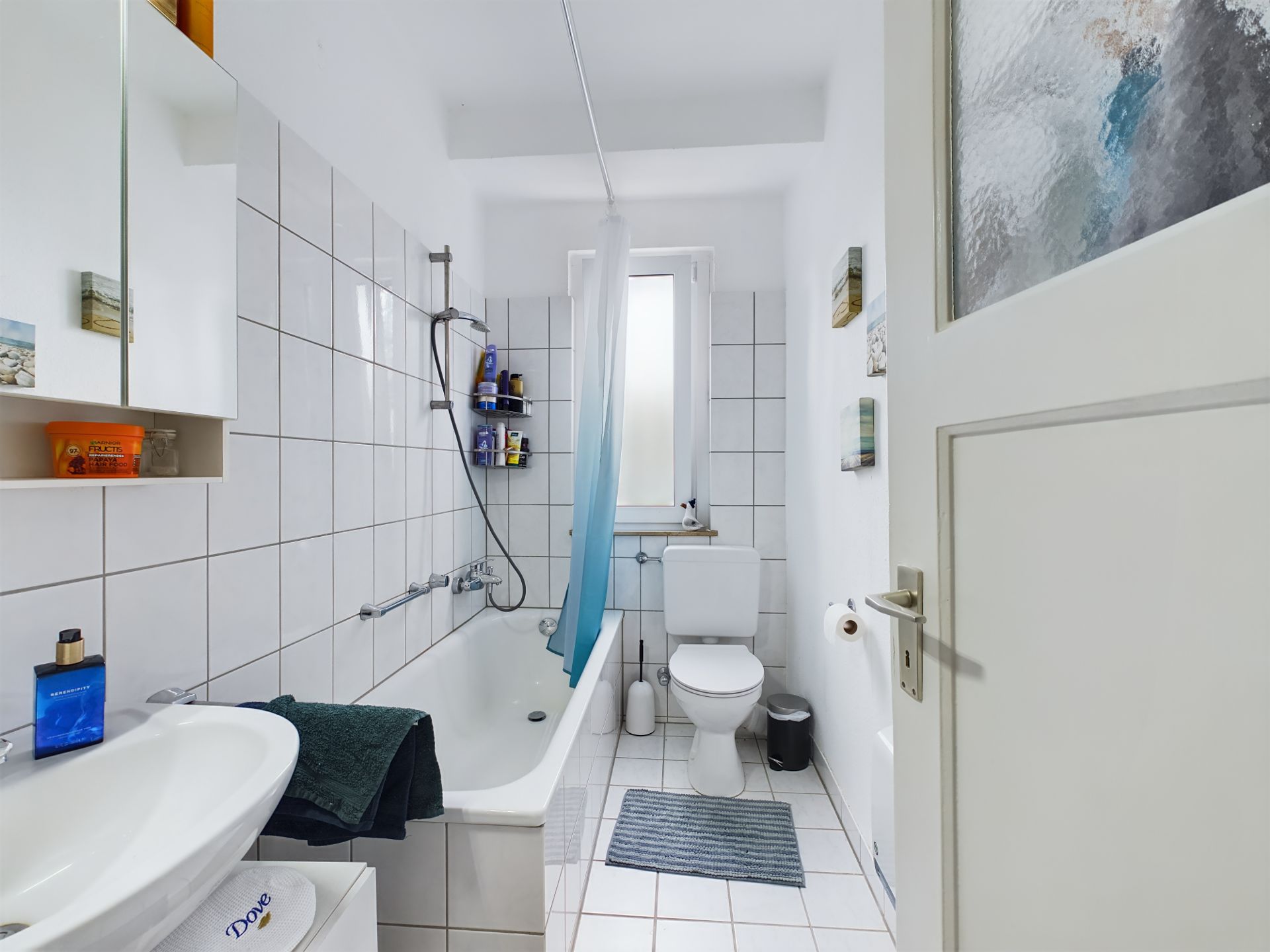 Helles Badezimmer mit Badewanne, Dusche, WC und Waschbecken, weiße Fliesen, Fenster und Deko an der Wand.