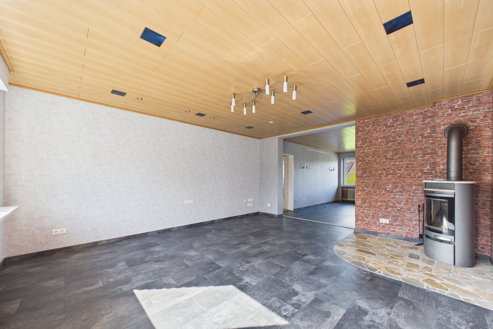 Leeres Wohnzimmer mit dunklem Boden, Holzdecke und Kamin an einer Ziegelwand.
