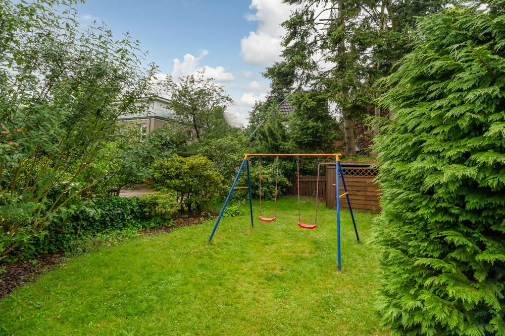 Garten mit zwei roten Schaukeln an einem blauen Metallgestell, umgeben von Bäumen und Büschen.