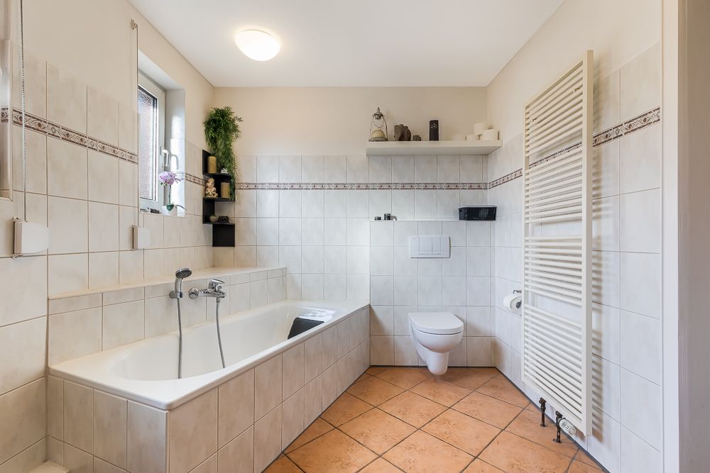 Helles Badezimmer mit Badewanne, Wandtoilette, Regal, Fenster und Handtuchheizkörper.