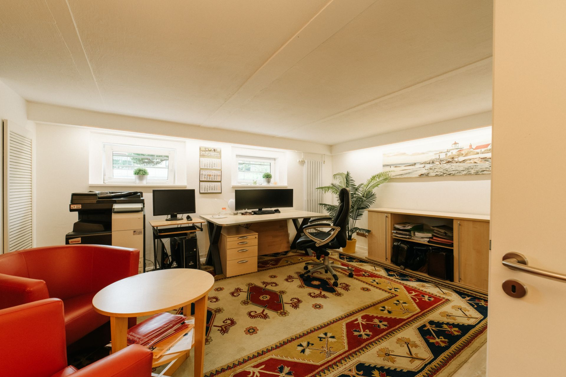 Helles Arbeitszimmer mit zwei Schreibtischen, Computern, rotem Sessel und großem Teppich.