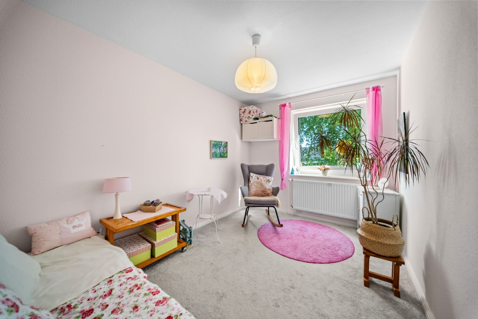Helles Kinderzimmer mit Einzelbett, Stuhl, Pflanze und Fenster mit pinken Vorhängen.