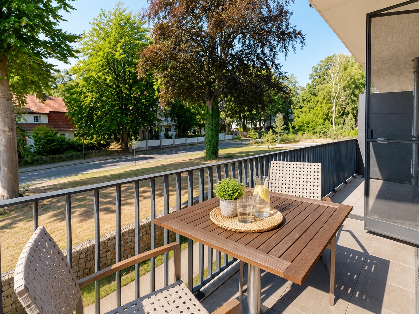 Balkon mit Holztisch, zwei Stühlen, einer Pflanze und Wasserkaraffe, Blick auf Bäume und Straße.