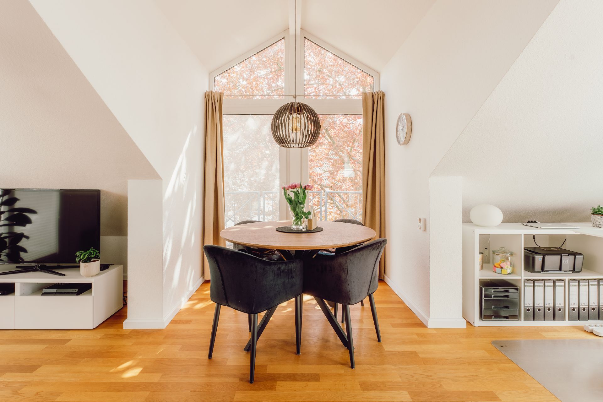 Essbereich mit rundem Tisch und vier Stühlen vor großem Fenster, daneben TV und Regal.