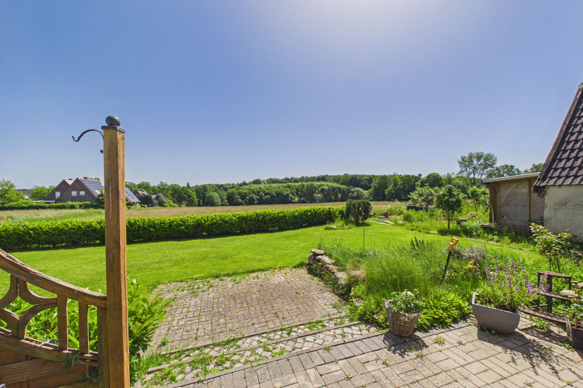 Gepflasterte Terrasse mit Blick auf Rasenfläche, Garten und offene Landschaft im Hintergrund.
