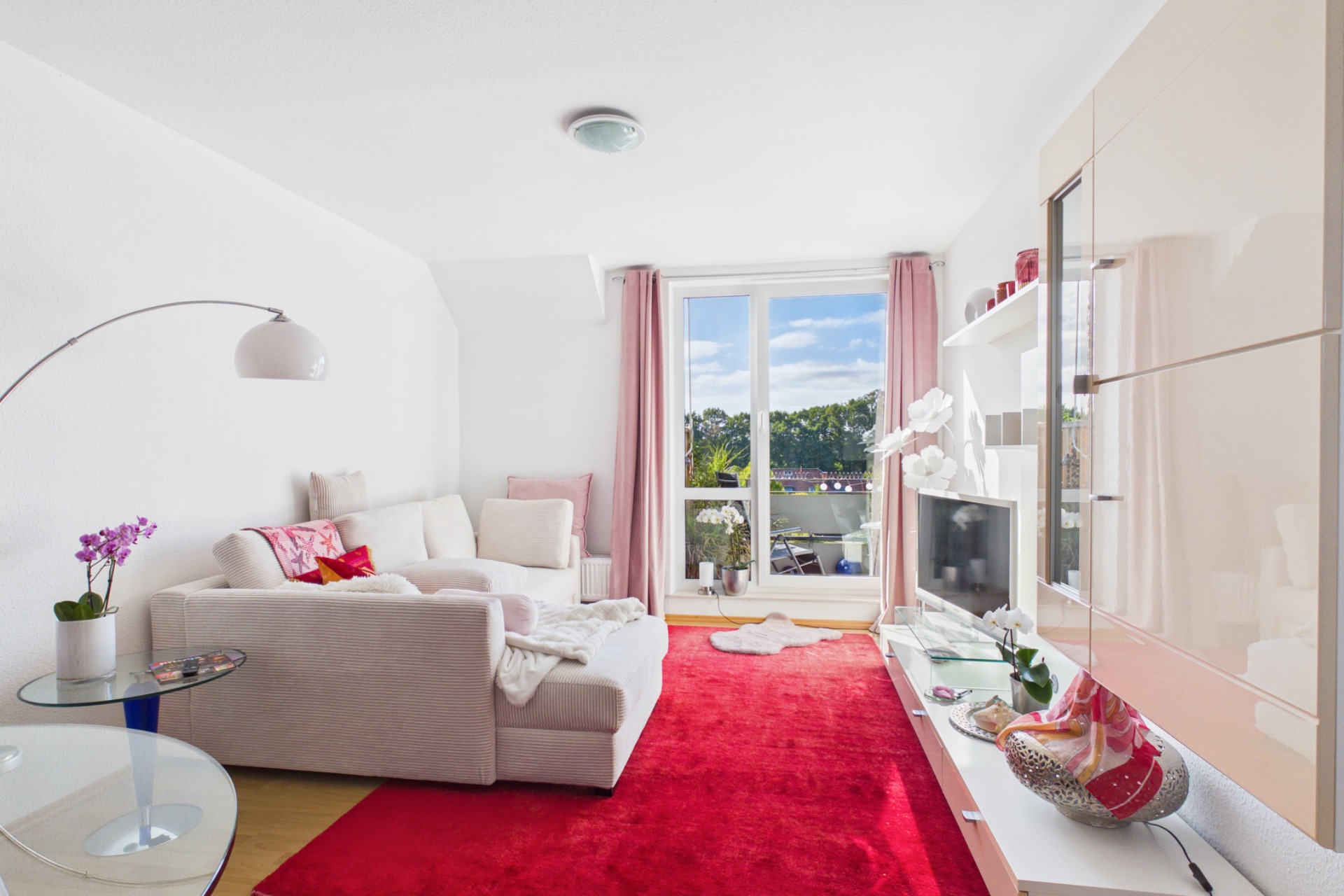 Helles Wohnzimmer mit Sofa, rotem Teppich, Glastisch und Zugang zu einem Balkon.
