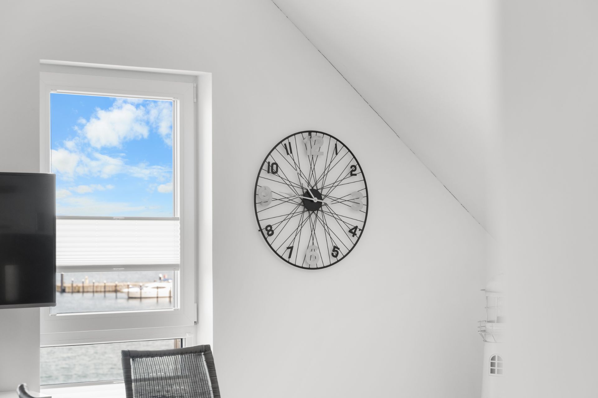 Helles Zimmer mit Wanduhr, Fenster mit Blick nach draußen und Stuhl im Vordergrund.