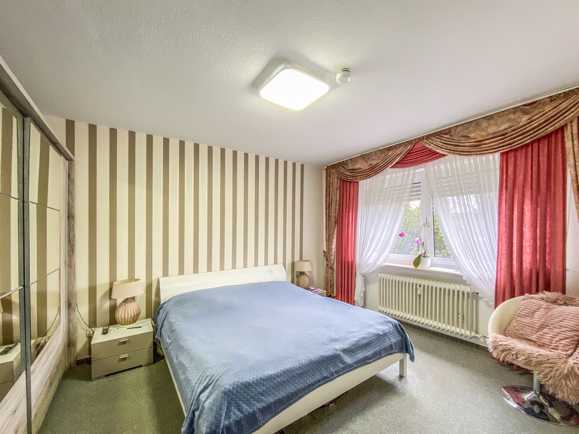 Schlafzimmer mit Doppelbett, Nachttischen, Spiegelkleiderschrank und Fenster mit Vorhängen.