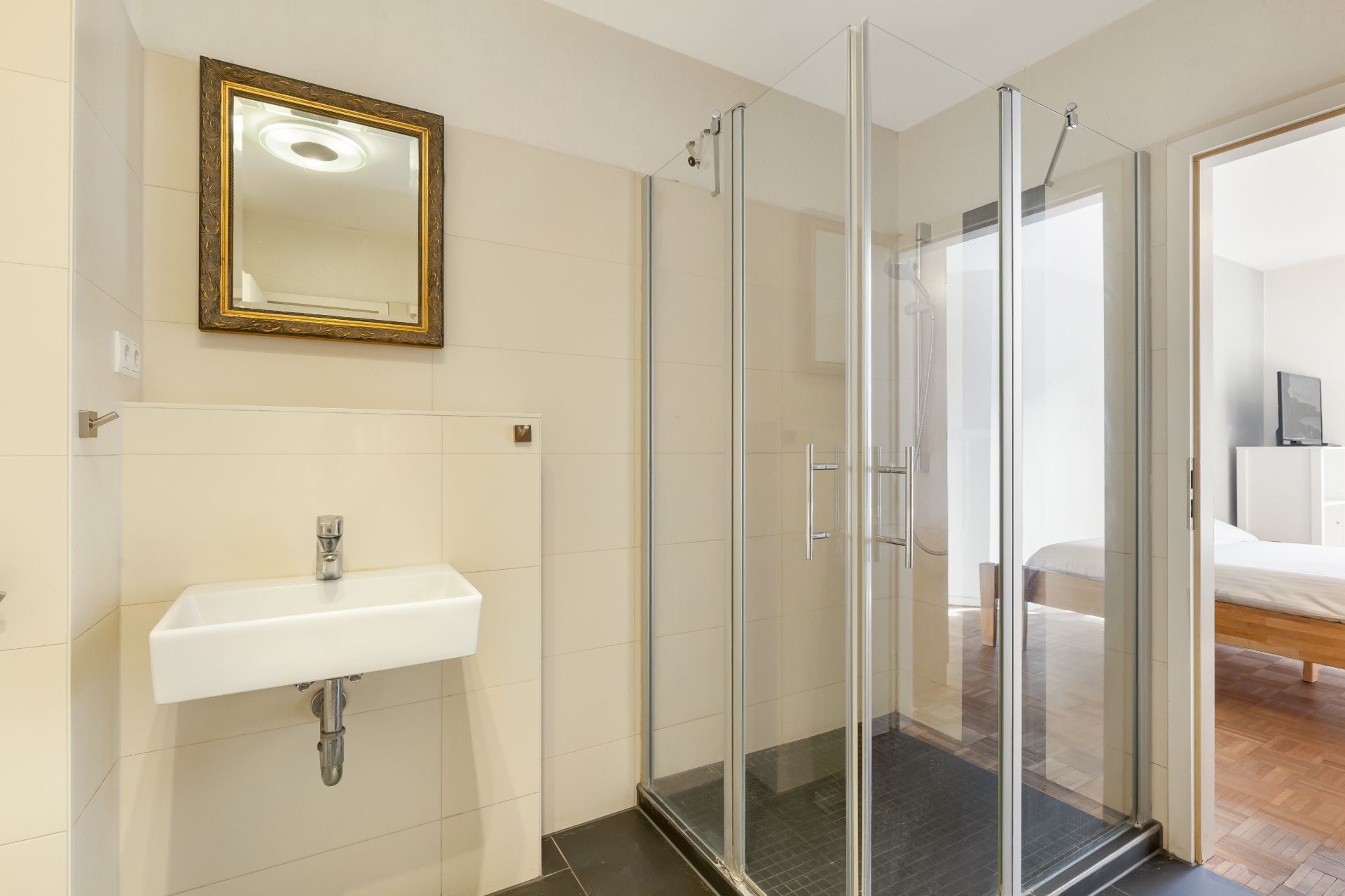 Modernes Badezimmer mit Eckdusche, Waschbecken und Spiegel, Tür führt in ein Schlafzimmer.