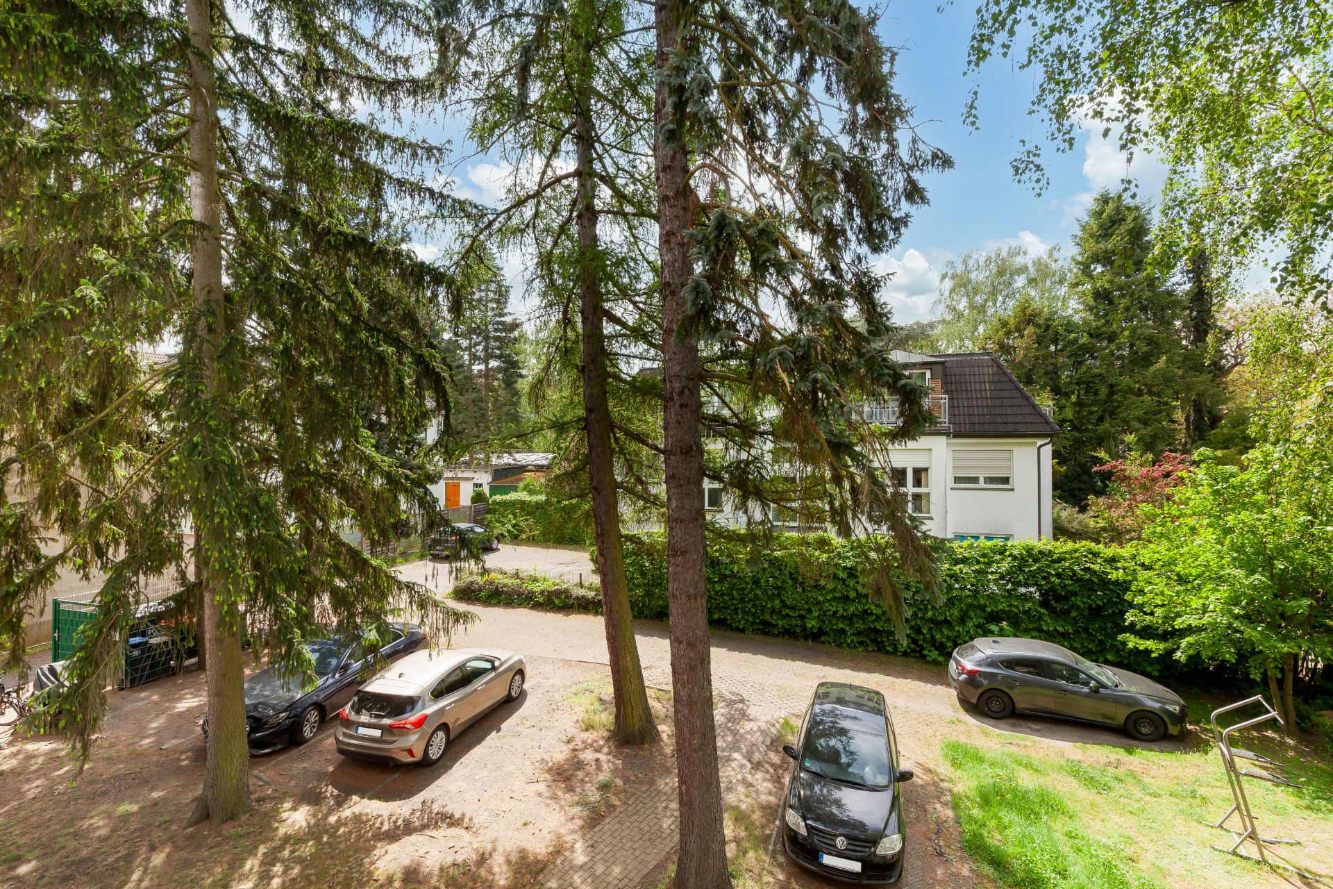 Parkplatz mit mehreren Autos und hohen Bäumen vor einem Wohnhaus bei Tageslicht.