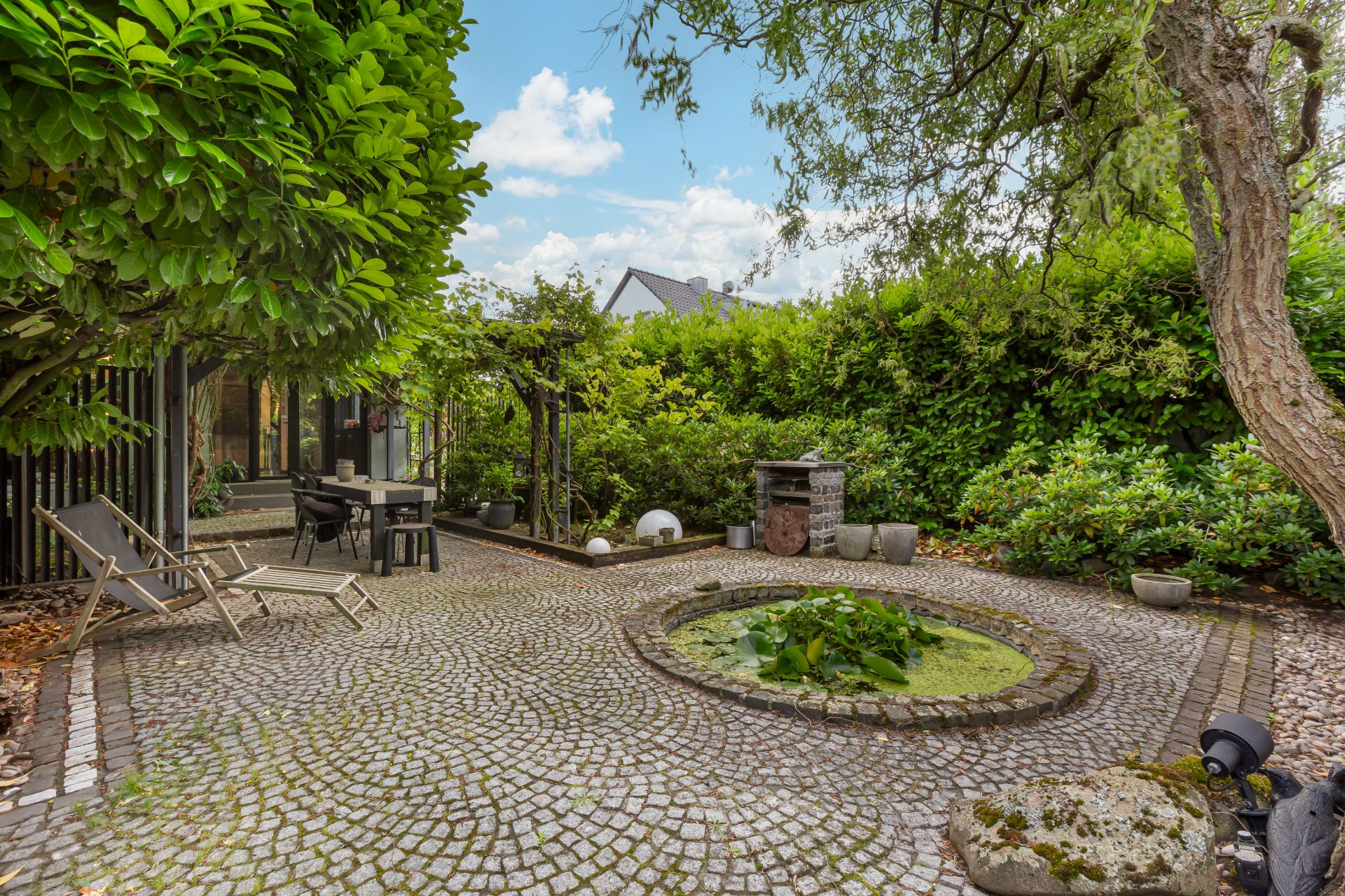 Gepflasterter Garten mit kleinem Teich, Gartenmöbeln und dichter Begrünung.