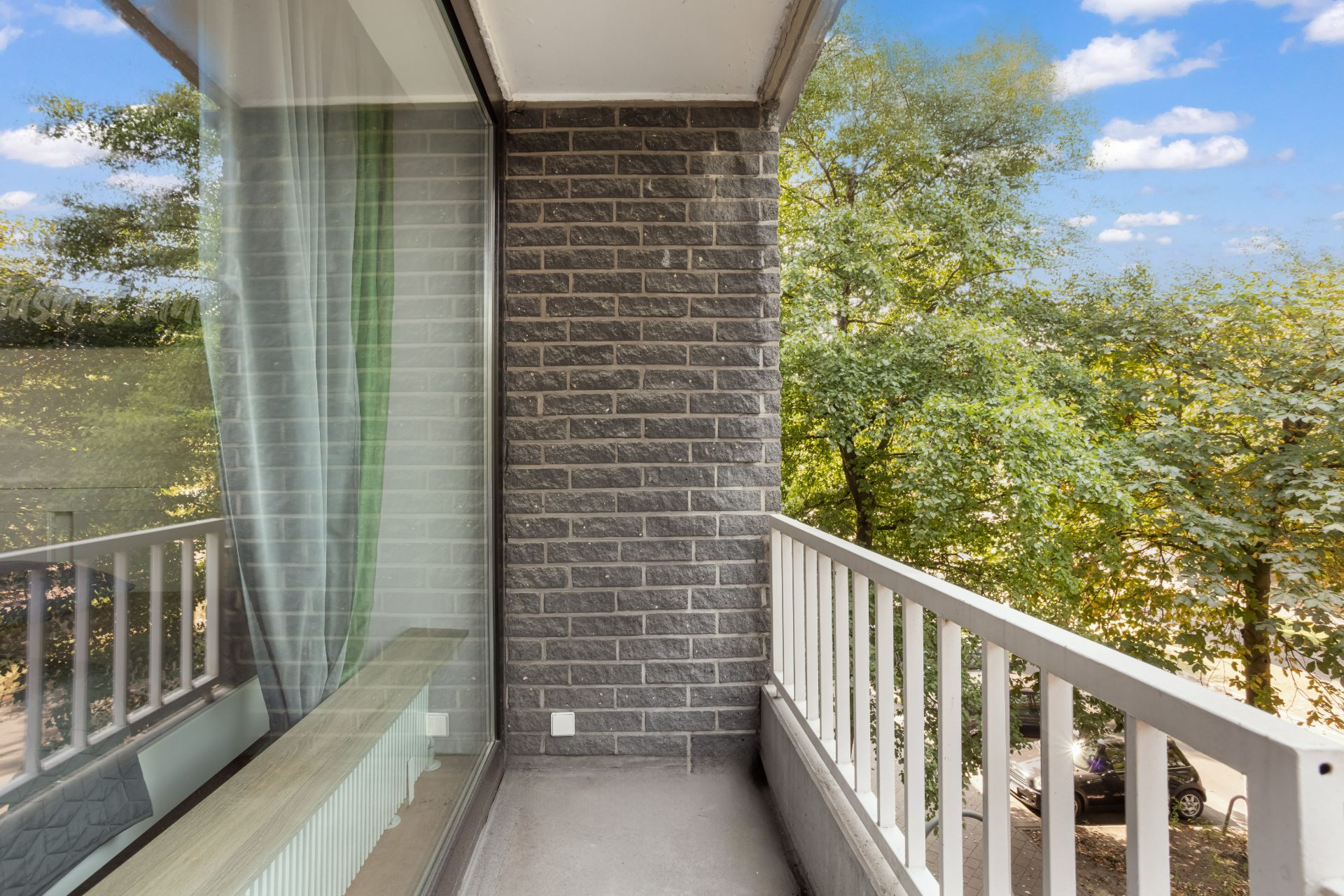 Kleiner Balkon mit grauer Backsteinwand und weißem Geländer, Blick auf grüne Bäume.