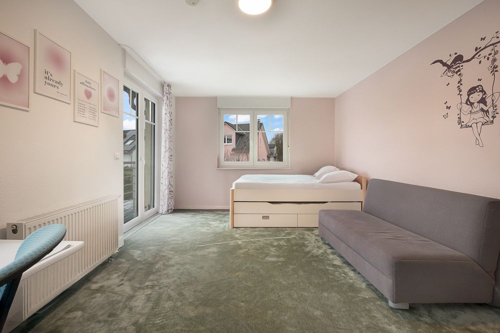 Helles Schlafzimmer mit Bett, grauem Sofa, Schreibtisch und Wanddeko auf rosa Wand.