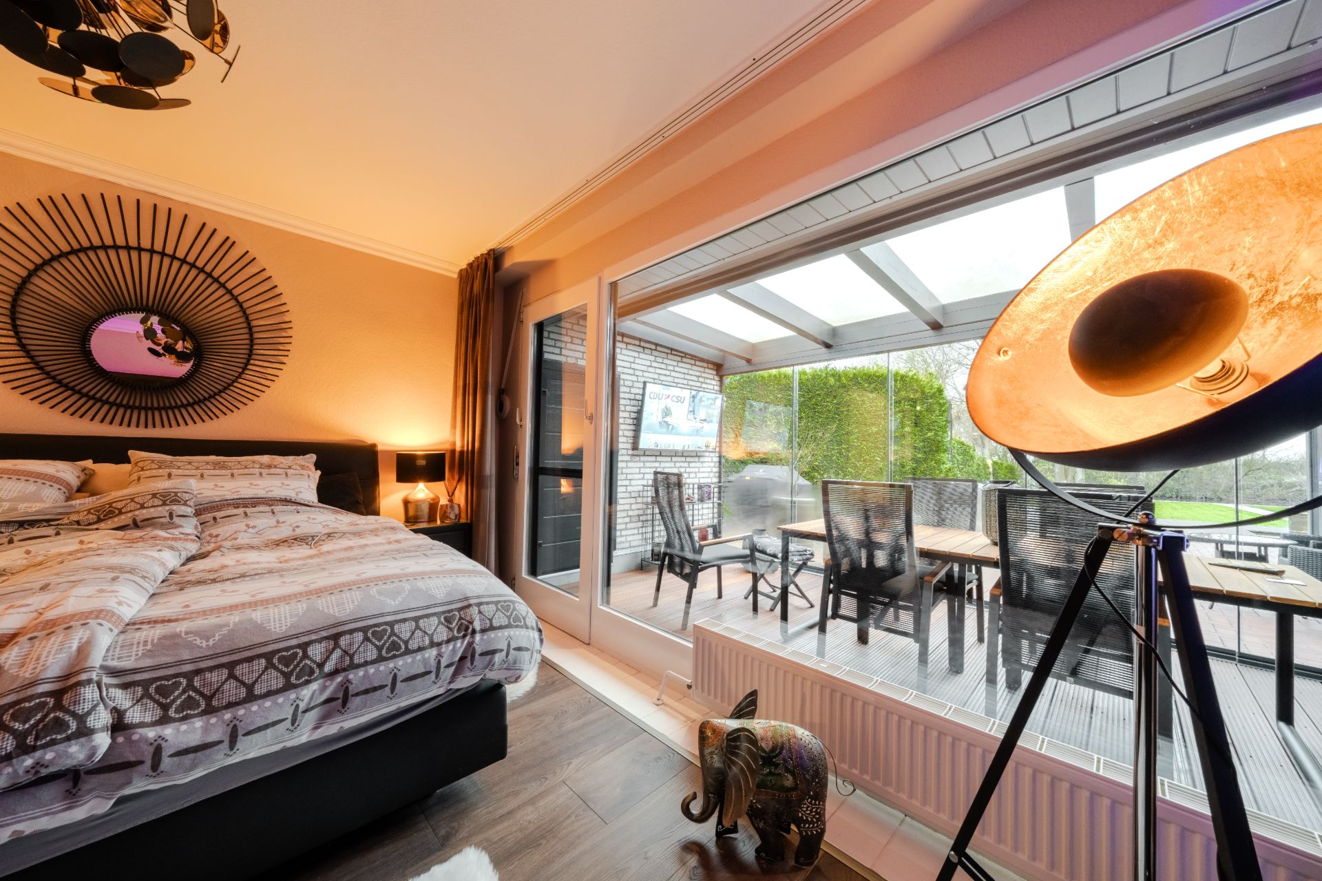 Schlafzimmer mit Doppelbett und großem Fenster zur überdachten Terrasse mit Esstisch und Stühlen.