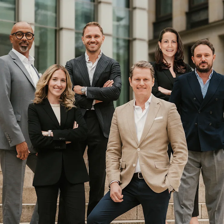 Sechs Erwachsene in Businesskleidung stehen und sitzen auf einer Treppe vor einem modernen Gebäude.