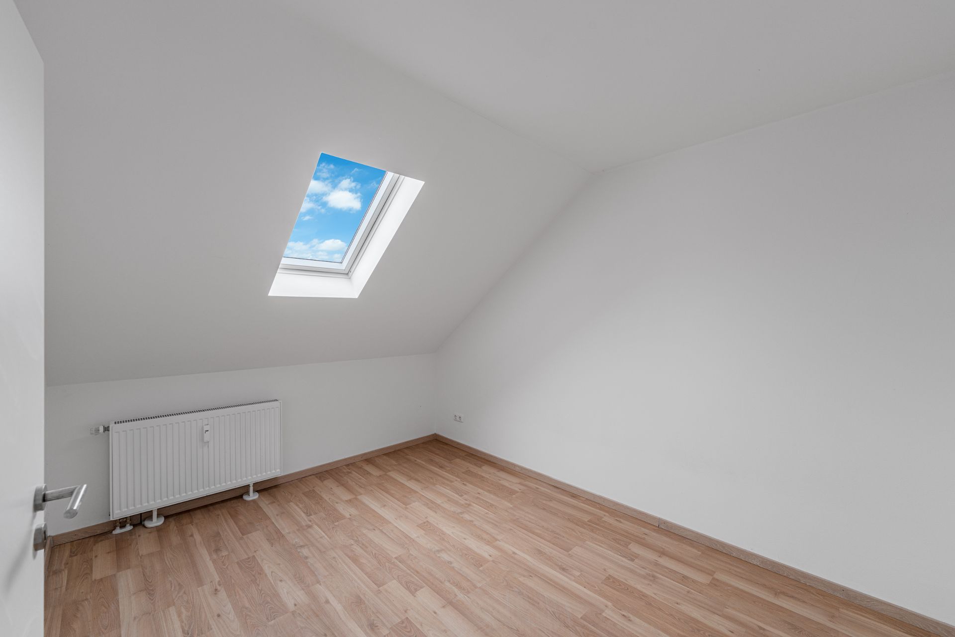 Leeres Zimmer mit schräger Decke, Dachfenster und Heizkörper, heller Holzboden.