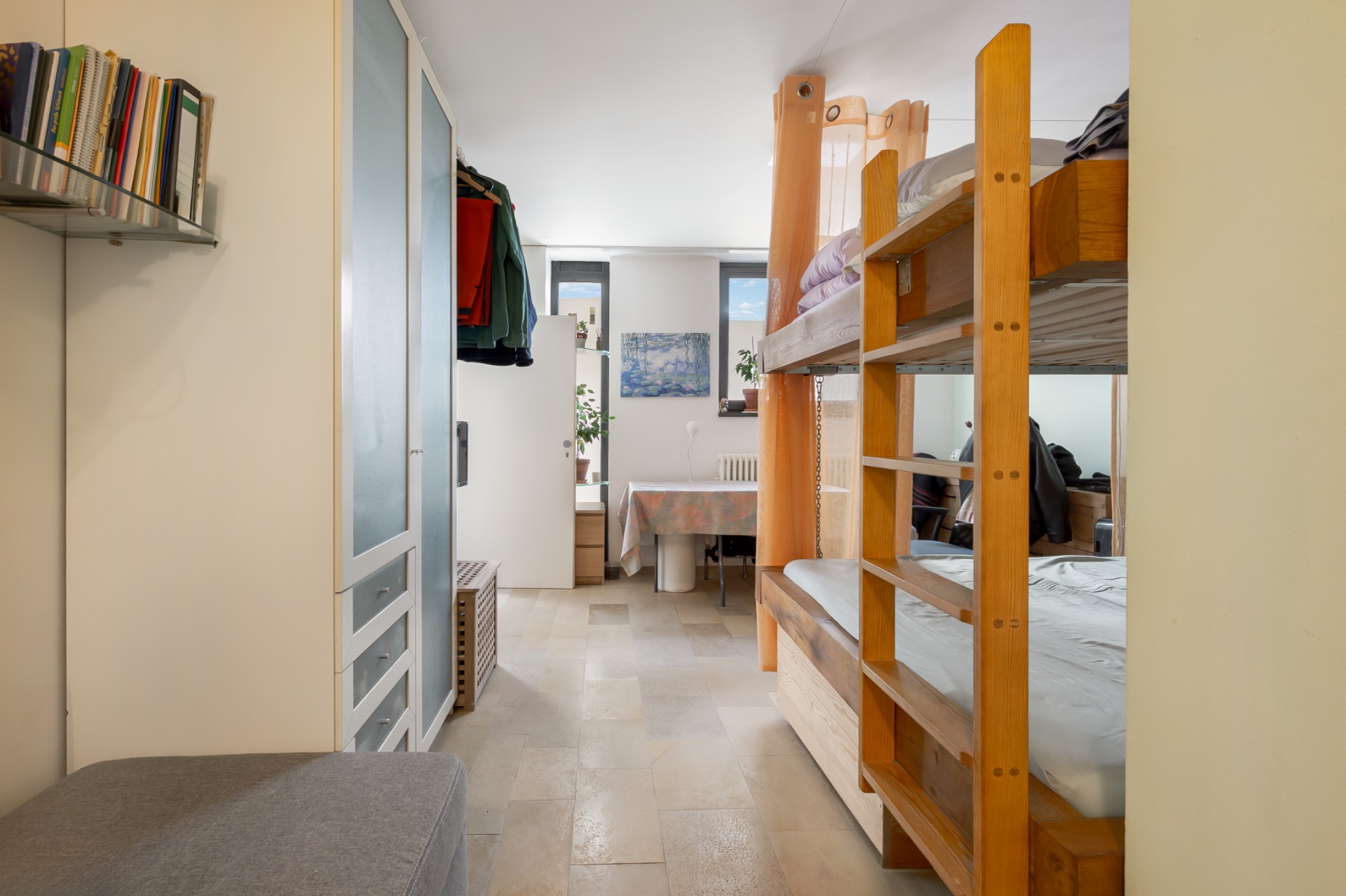 Zimmer mit Etagenbett, Schrank, Tisch am Fenster und Bücherregal an der Wand.