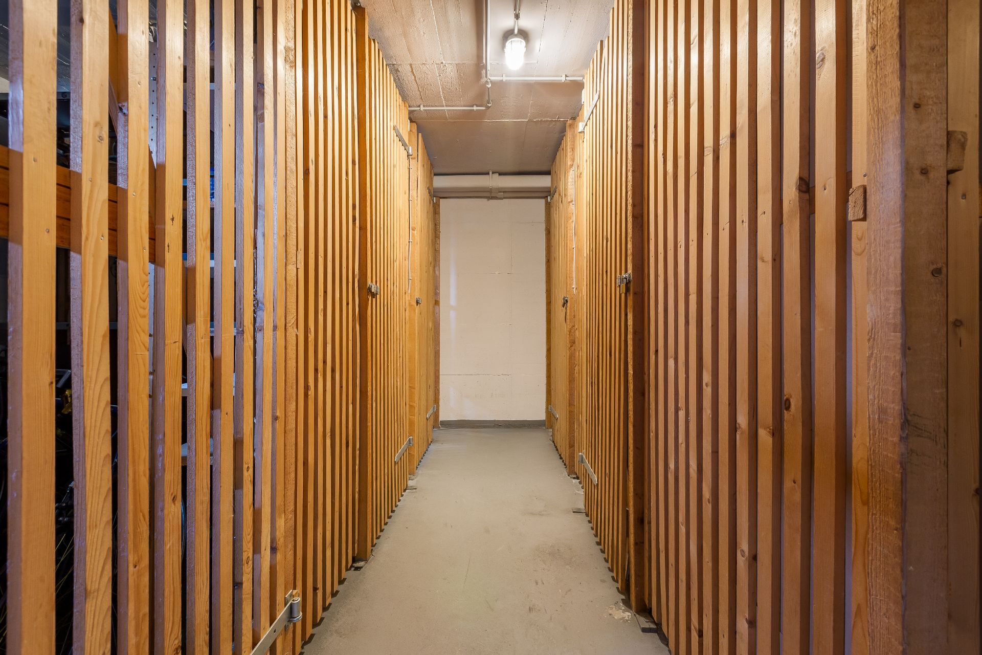 Gang mit Abteilen aus Holzlatten in einem Kellerraum, Deckenlampe leuchtet.
