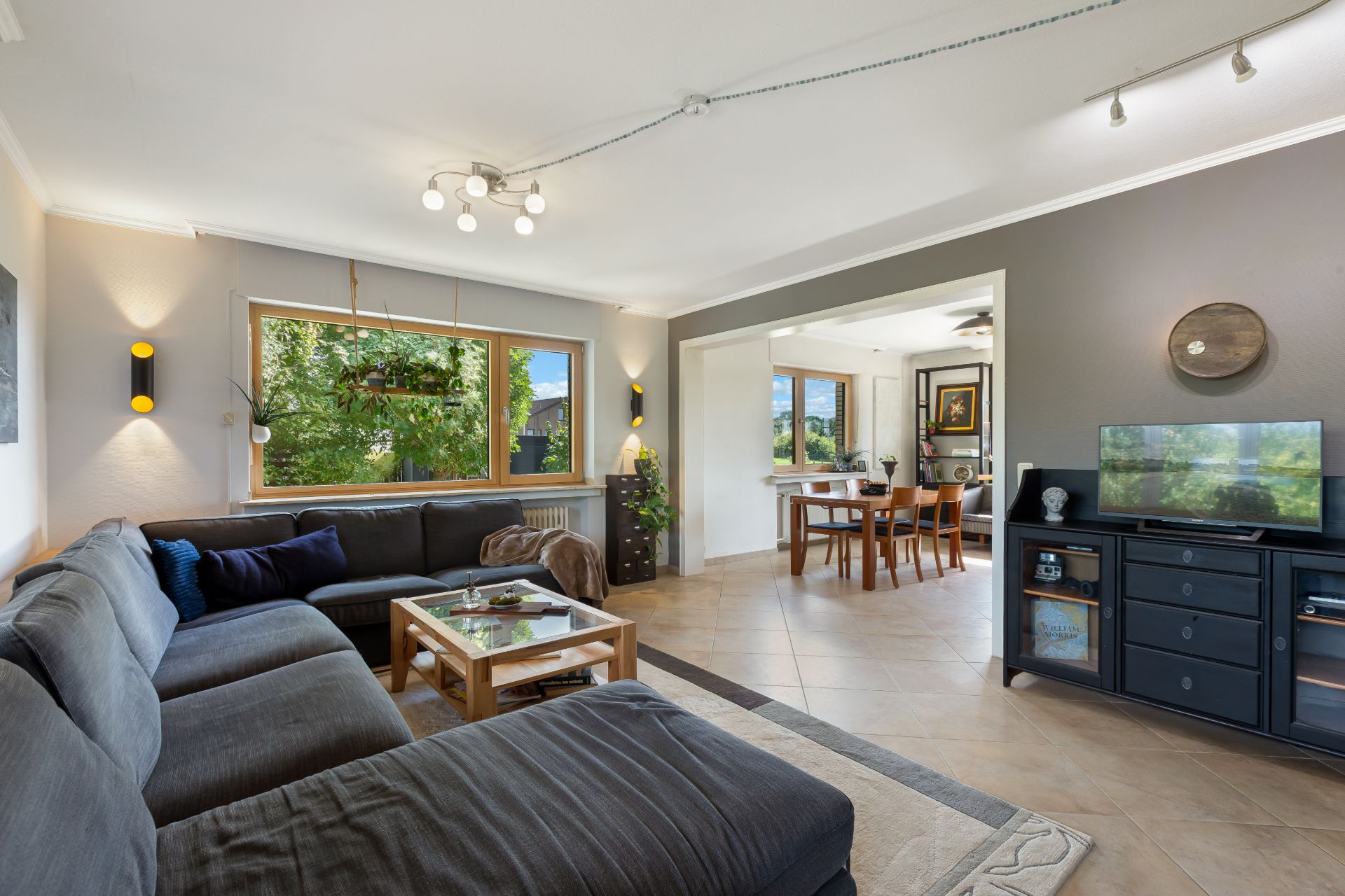 Modernes Wohnzimmer mit großer grauer Eckcouch, Fernseher und Essbereich im Hintergrund.