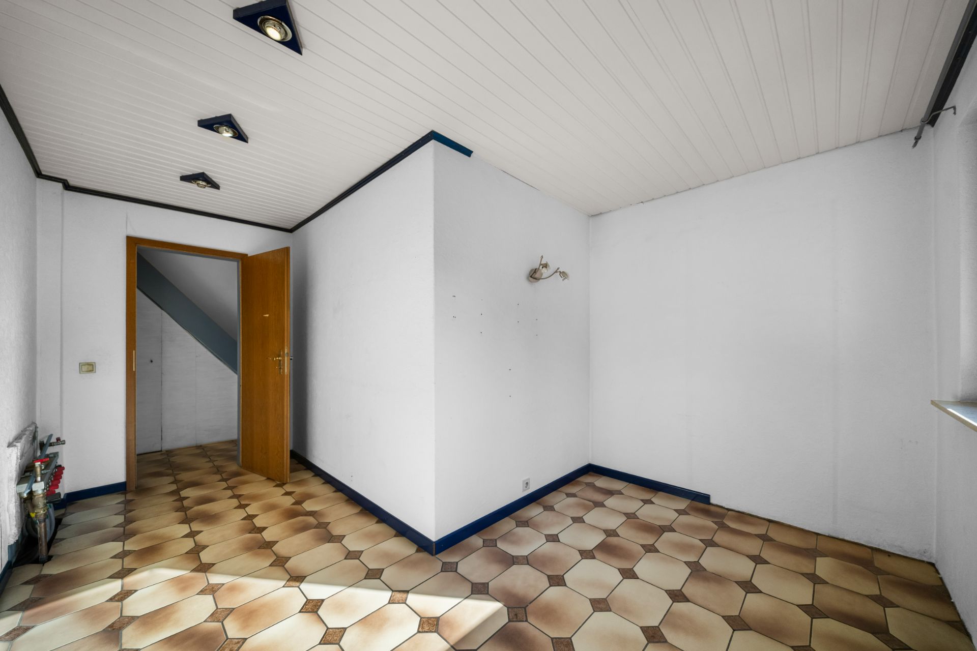 Leerer Raum mit gefliestem Boden, weißer Decke und offenstehender Holztür.