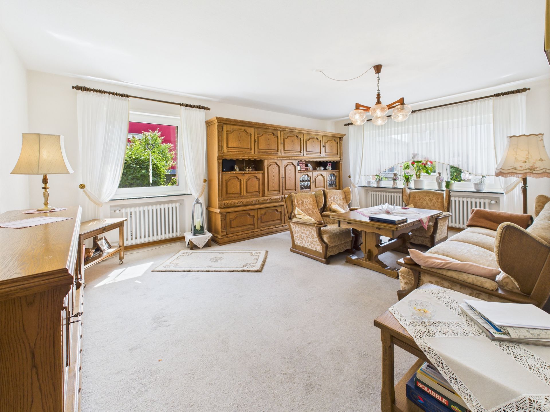 Helles Wohnzimmer mit Holzmöbeln, Sofa, Sessel, großem Schrank und mehreren Fenstern mit weißen Vorhängen.