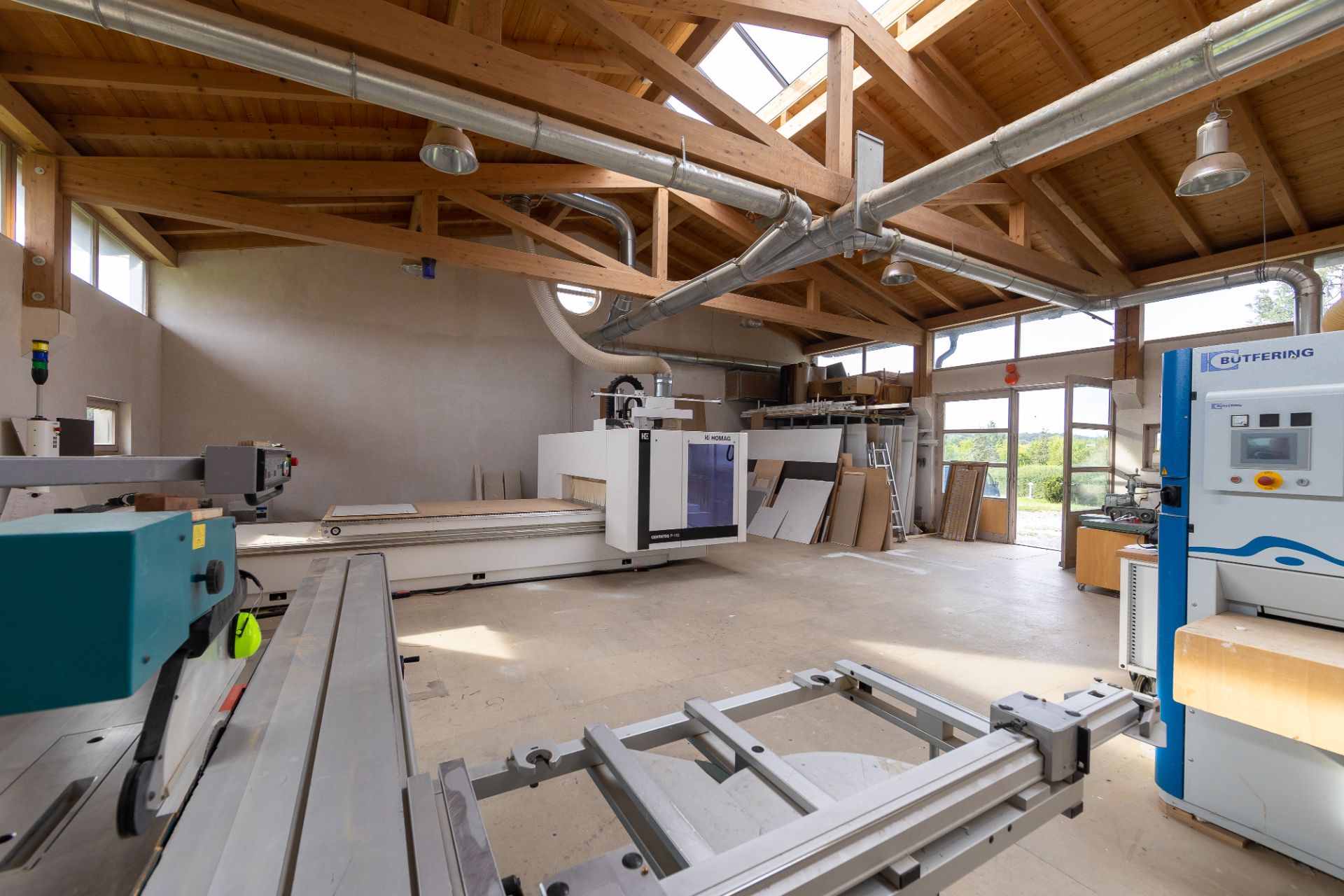 Helle Werkstatt mit Holzbalkendecke, Maschinen und großen Fenstern.