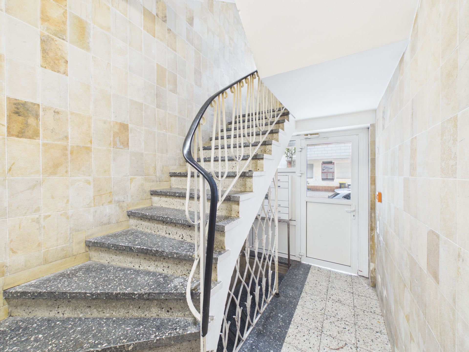 Helle Treppenhaus mit Steintreppe, Geländer und Hauseingangstür mit Briefkästen.