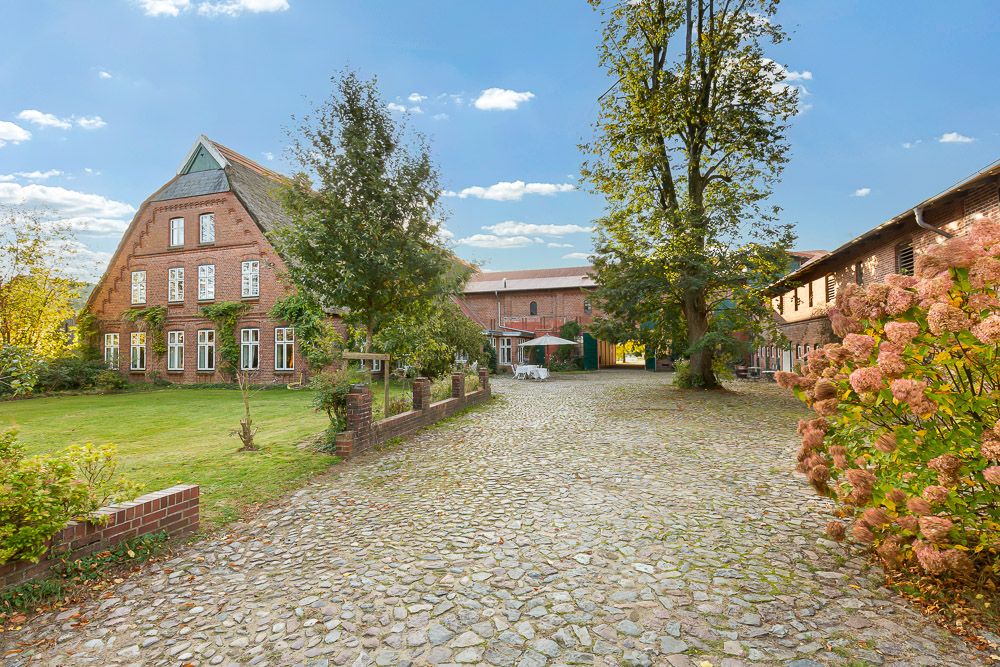 Gepflasterter Hof mit Backsteingebäuden, Bäumen und Rasenfläche, sonniges Wetter.