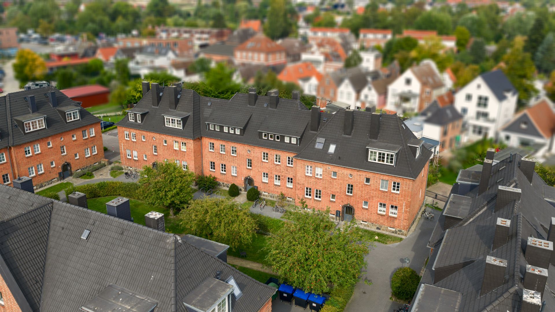 Mehrere rote Wohngebäude mit dunklen Dächern und grünem Innenhof aus der Vogelperspektive.