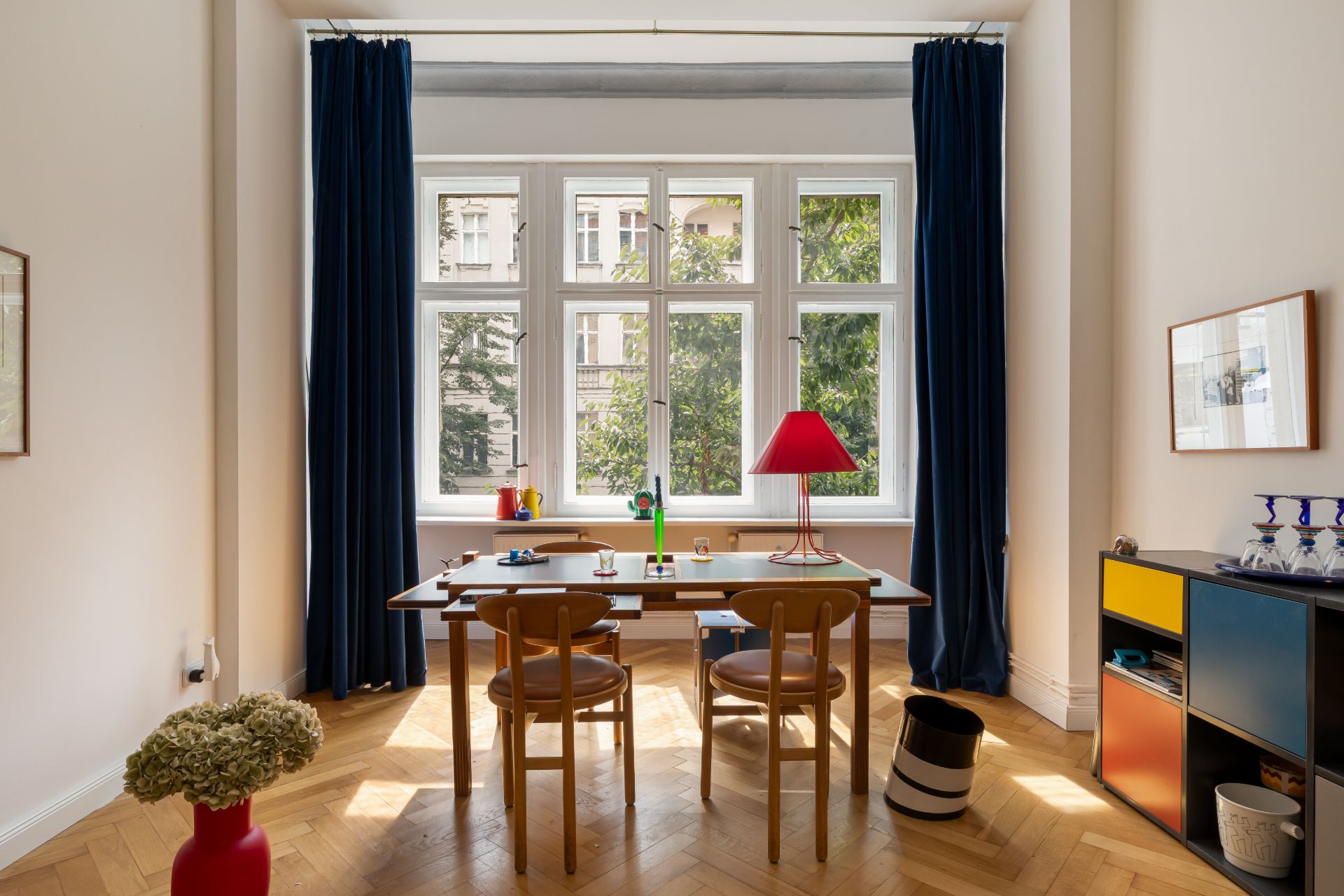 Heller Raum mit großem Fenster, Holztisch, drei Stühlen und buntem Regal an der Seite.