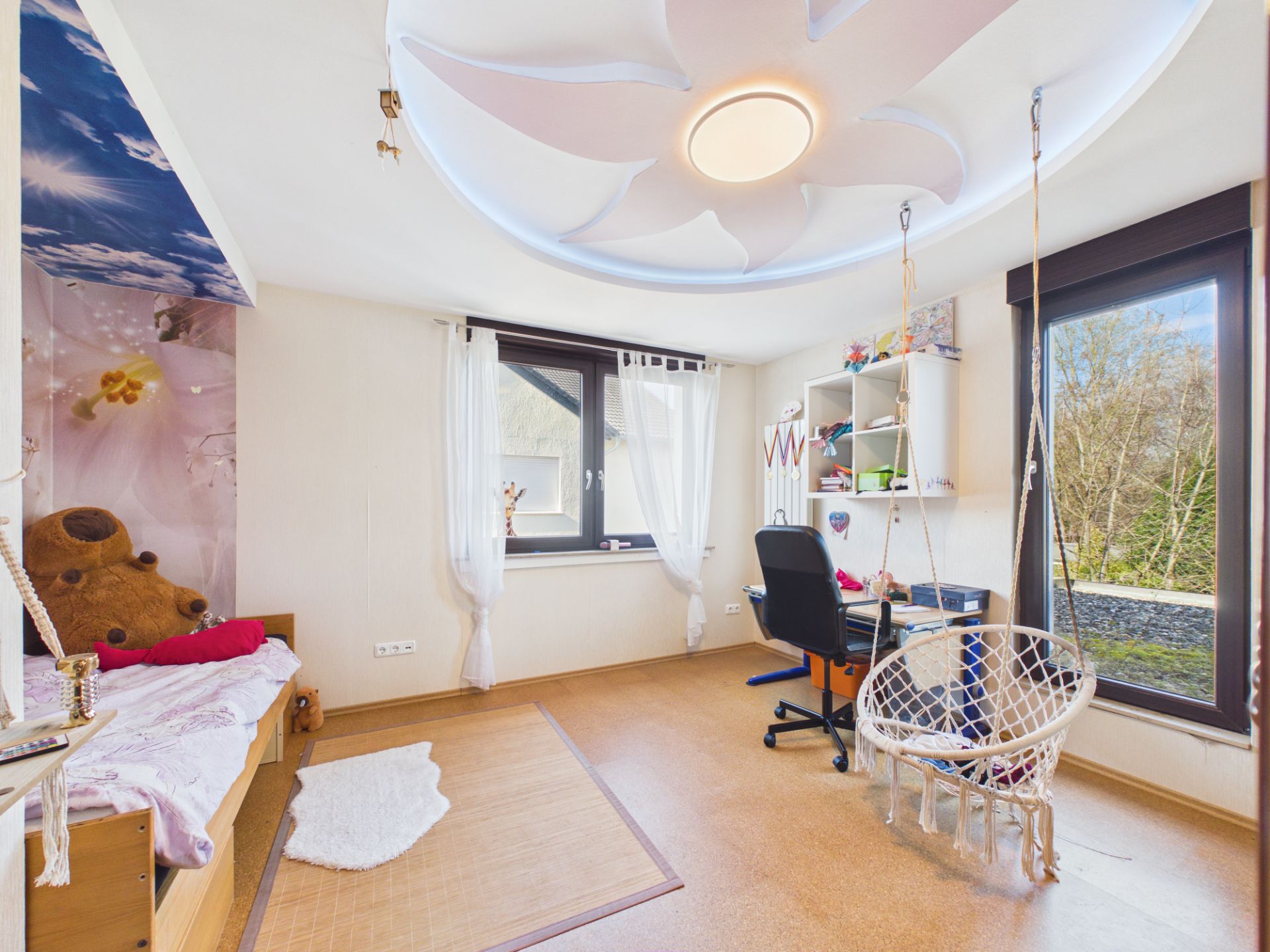 Helles Kinderzimmer mit Schreibtisch, Hängesessel, Bett und großen Fenstern.