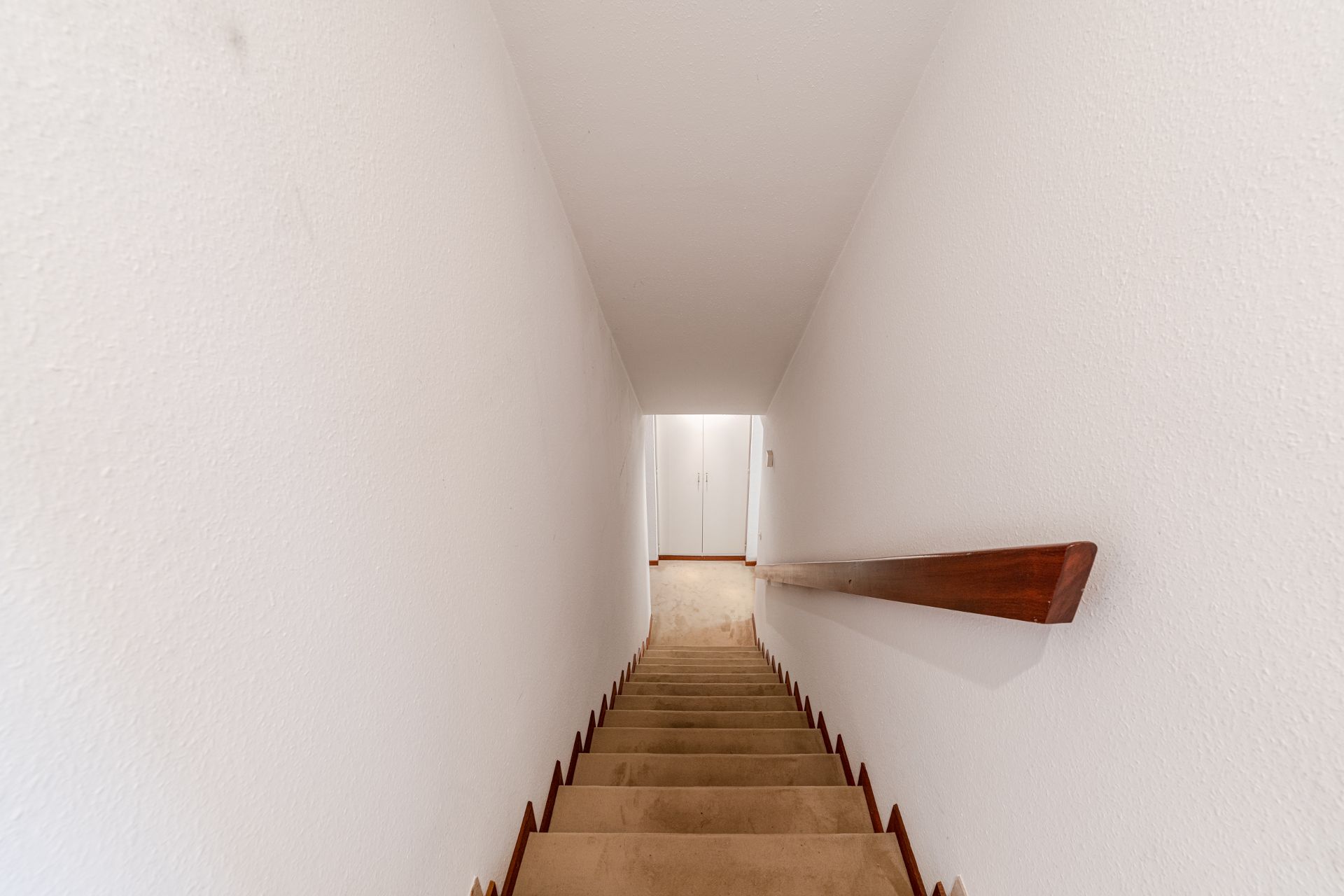 Beige Treppe mit Holzgeländer zwischen weißen Wänden, am Ende eine weiße Tür.