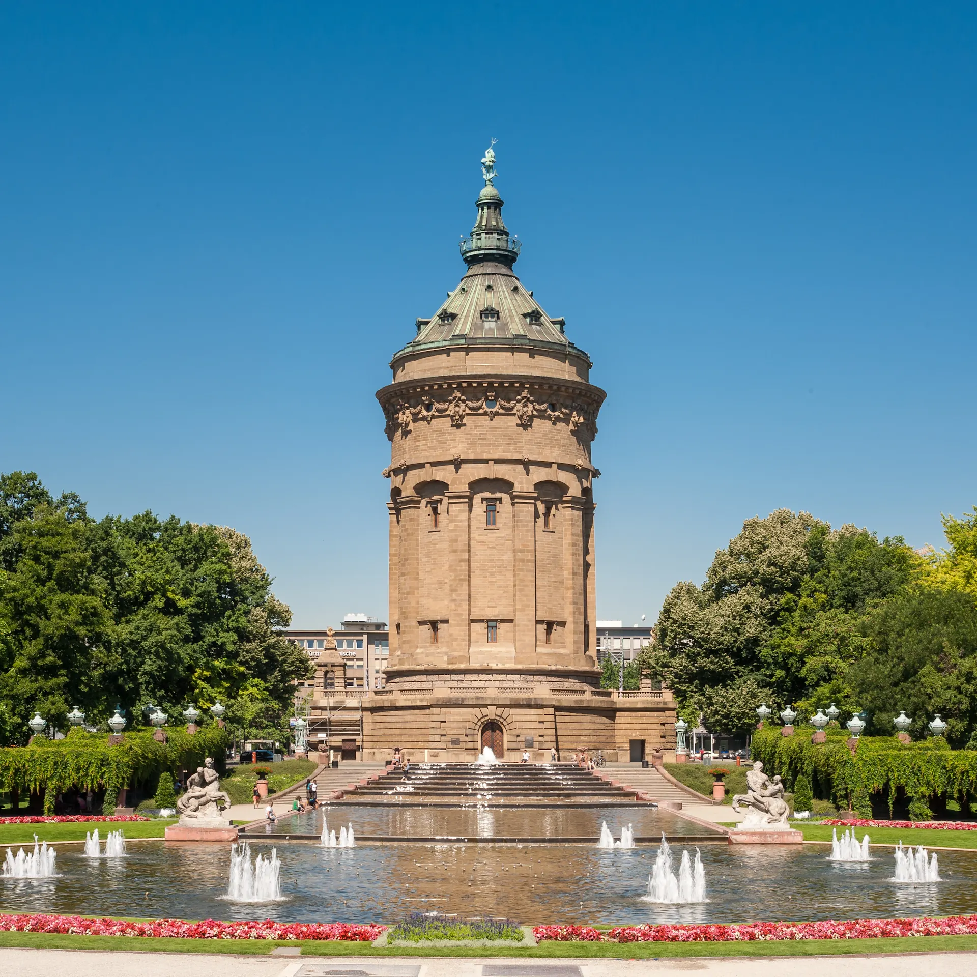 Ein großer, runder Turm vor einem Springbrunnen in einer grünen Parkanlage.