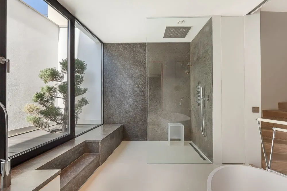Modernes Badezimmer mit großer Glasdusche, Sitzbank am Fenster und Blick auf einen kleinen Baum draußen.