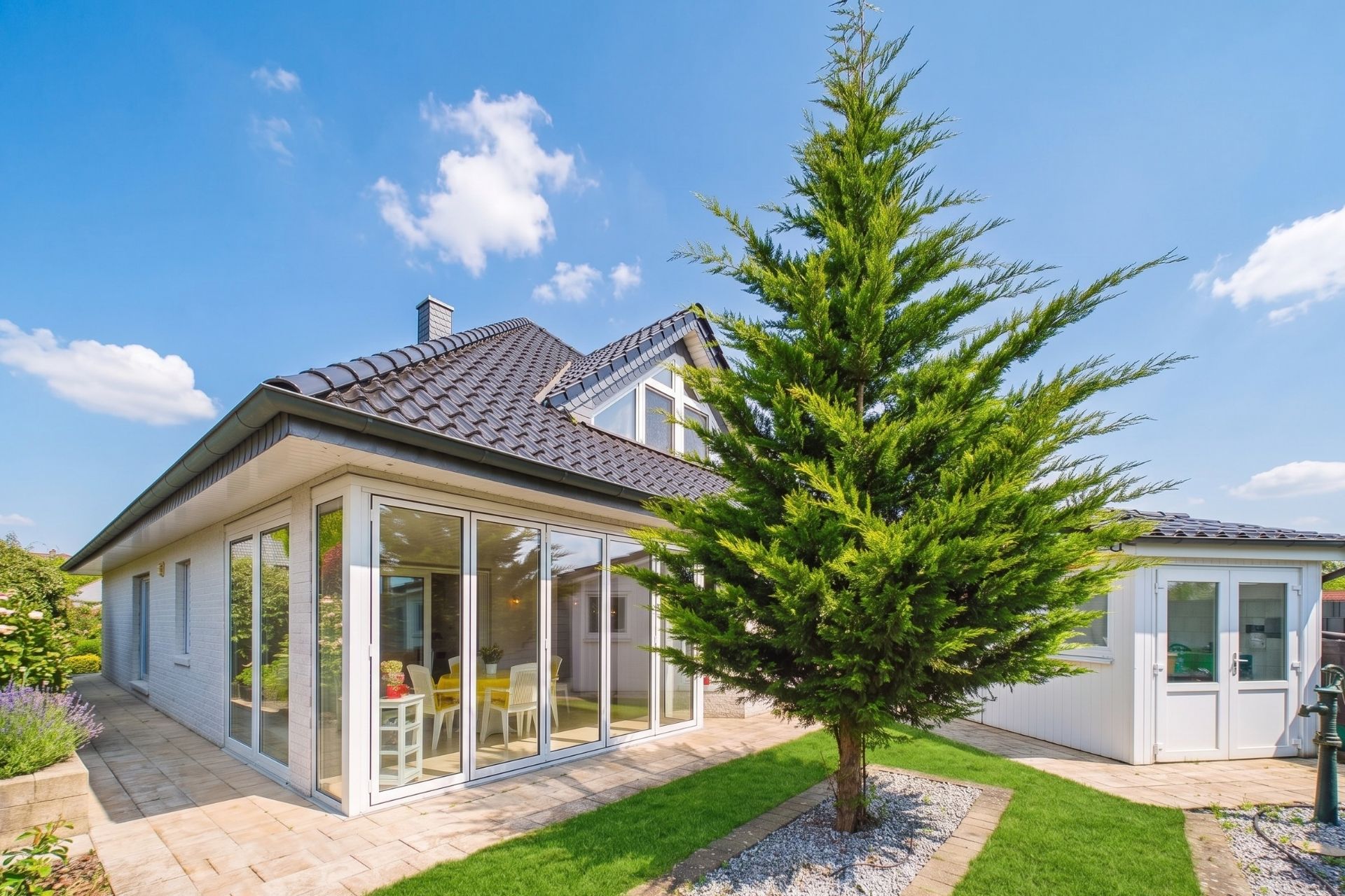 Modernes Einfamilienhaus mit Wintergarten und großem Baum im Garten bei sonnigem Wetter.