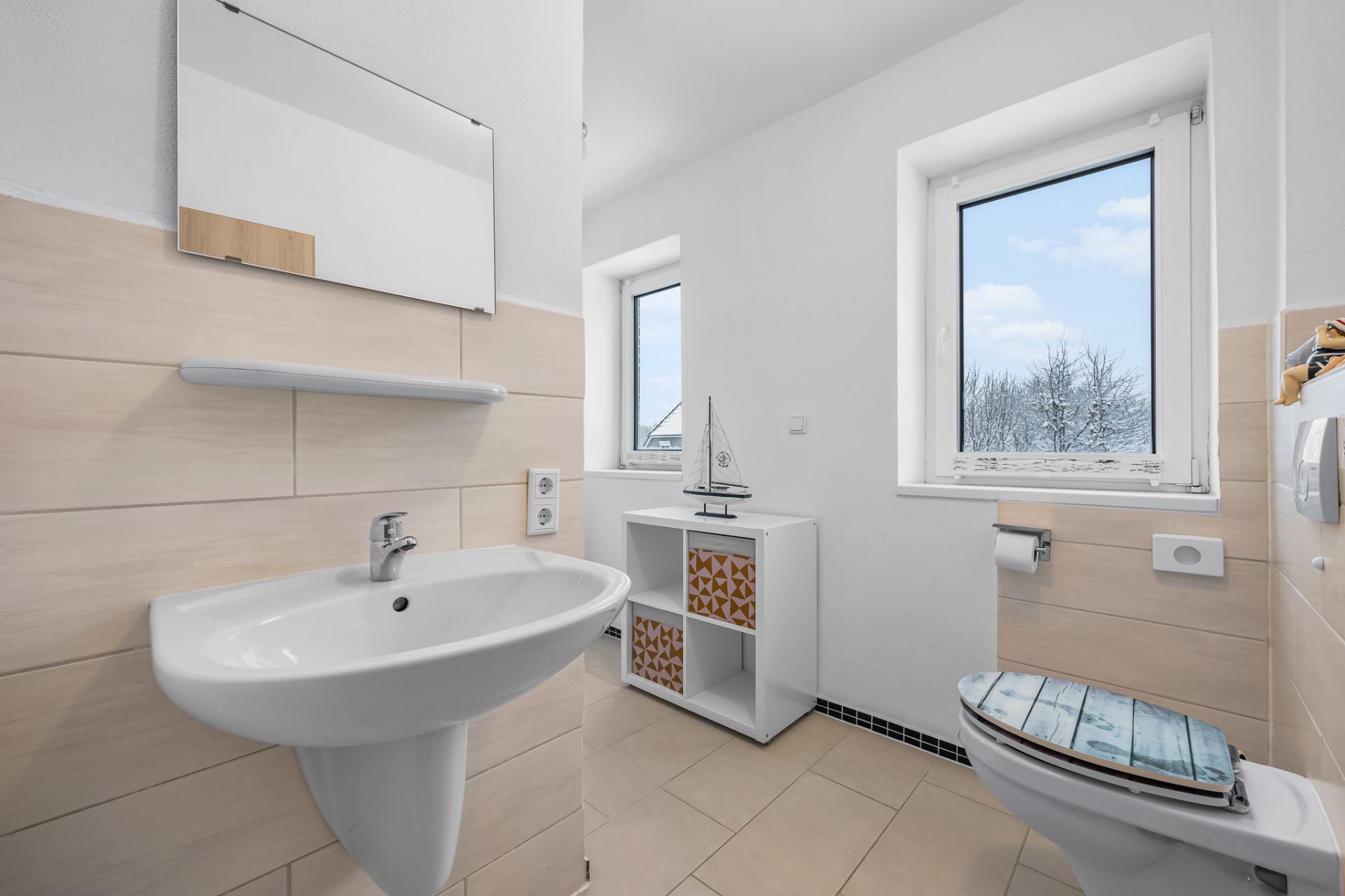 Helles Badezimmer mit Waschbecken, Spiegel, Toilette und Regal vor zwei Fenstern.