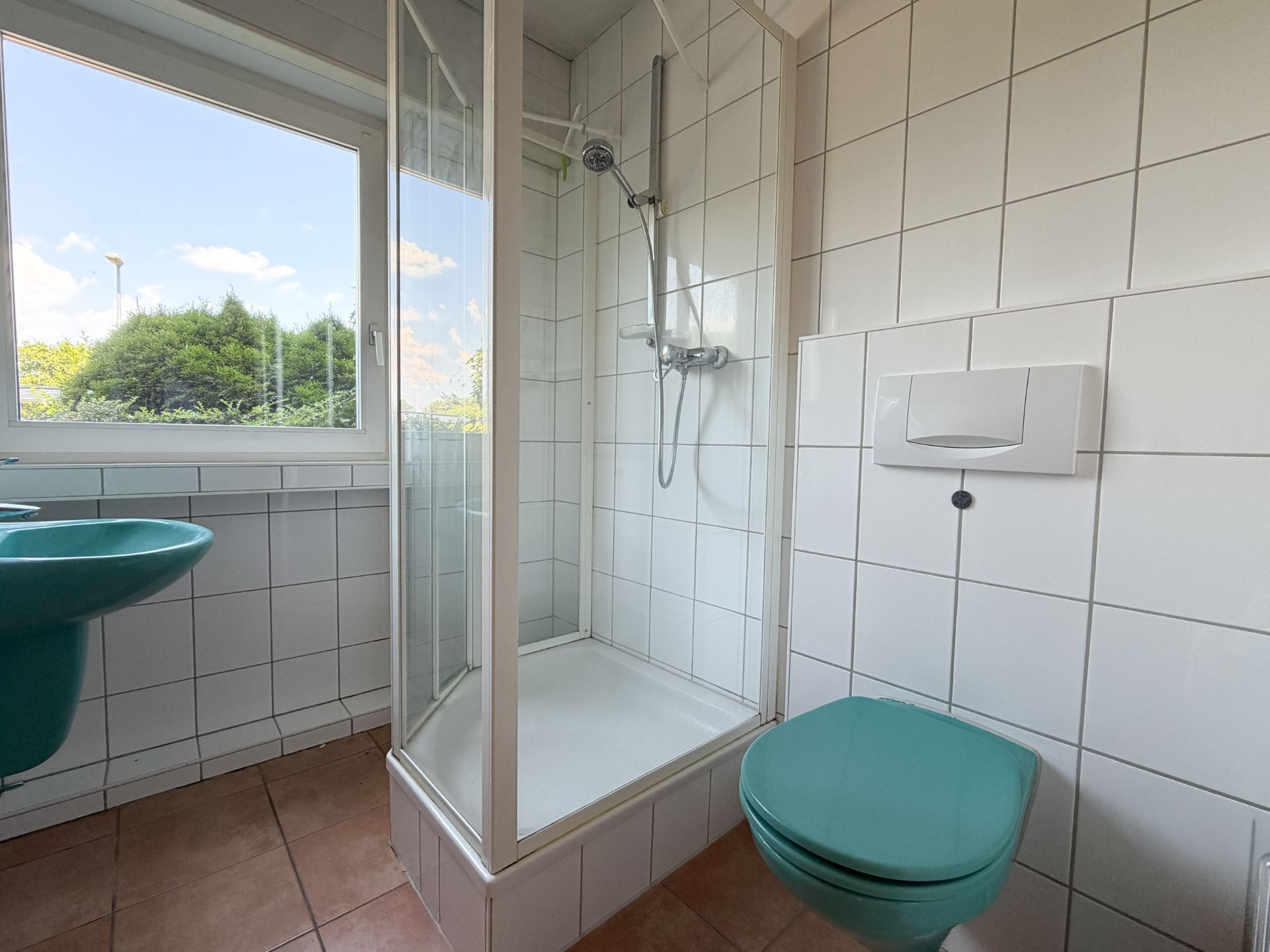 Kleines Bad mit Dusche, Fenster, grünem Waschbecken und grünem WC auf weißen Fliesen.