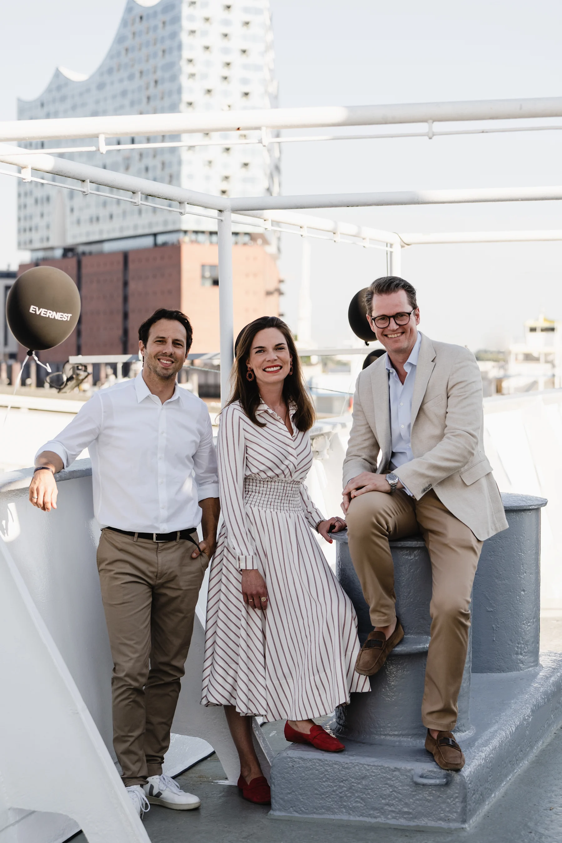 Drei lächelnde Personen stehen auf einem Schiff, im Hintergrund moderne Gebäude.