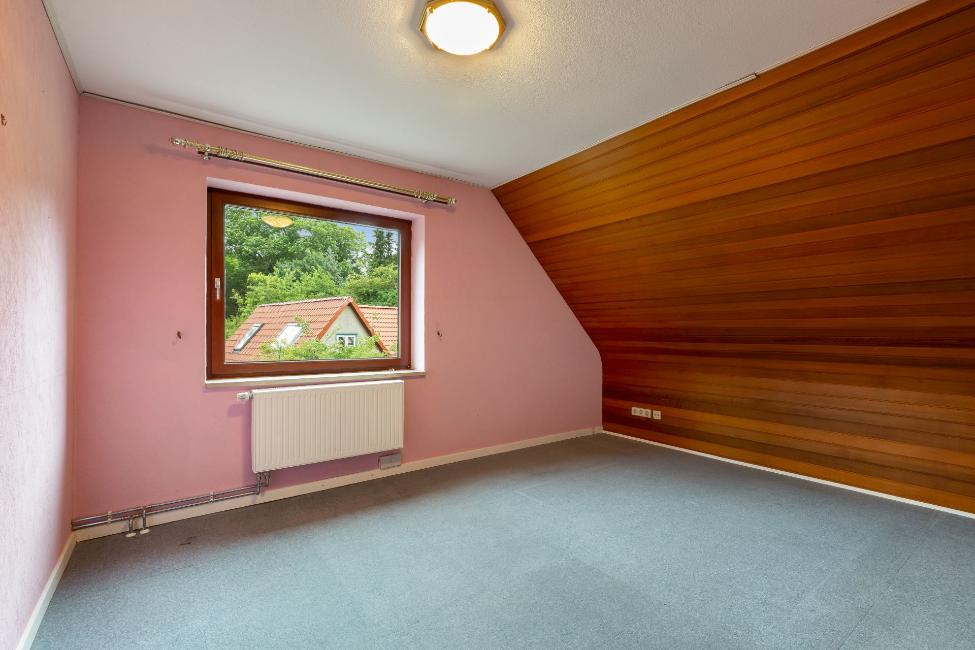 Leeres Zimmer mit rosa Wänden, Holzverkleidung und Fenster mit Blick auf Häuserdächer.