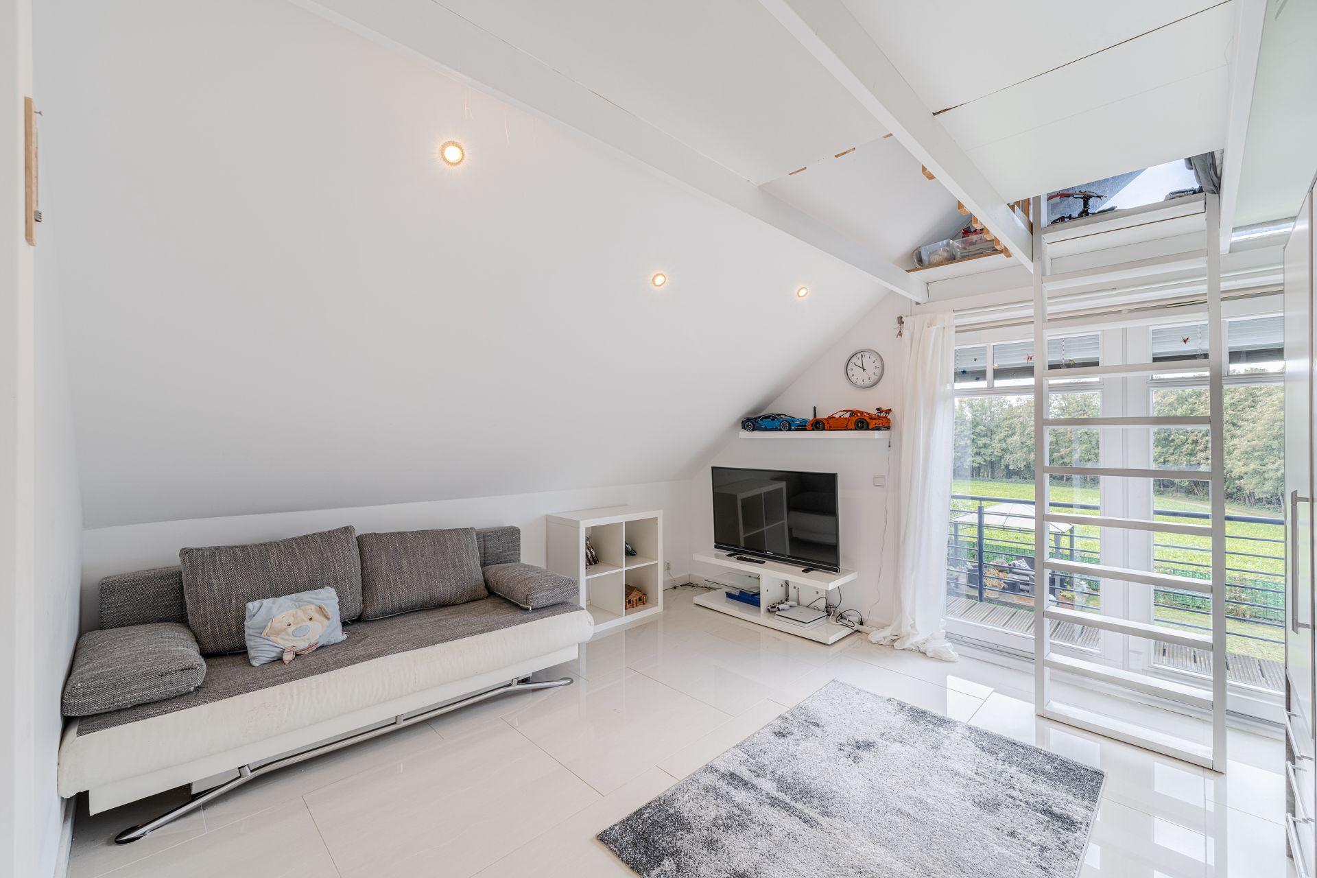 Helles Wohnzimmer mit grauem Sofa, Fernseher, Teppich und großer Fensterfront mit Blick ins Grüne.