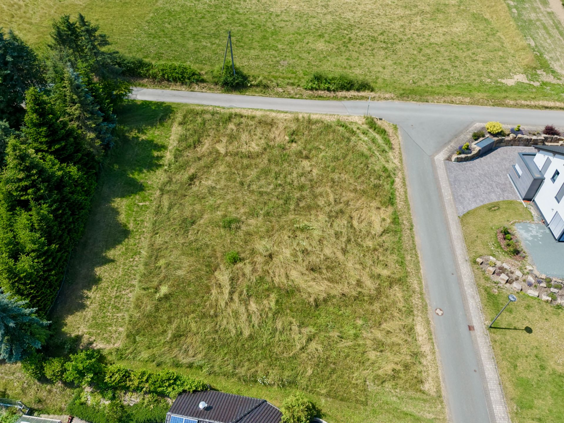 Luftaufnahme eines unbebauten, grasbewachsenen Grundstücks an einer Straßenecke, neben Wohnhäusern.
