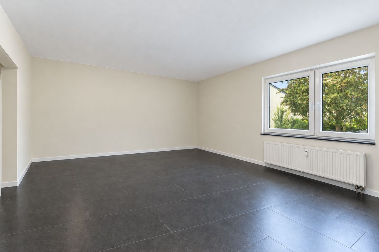 Leerer Raum mit hellen Wänden, großem Fenster und dunklem Fliesenboden.
