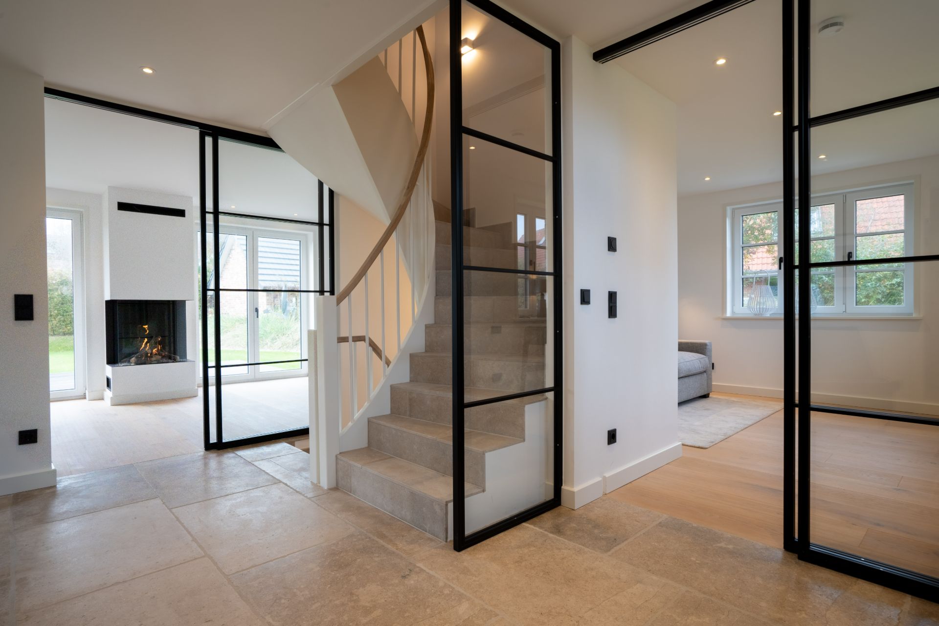 Heller Flur mit modernen Glastüren, Treppe und Blick in Wohnzimmer mit Sofa und Kamin.