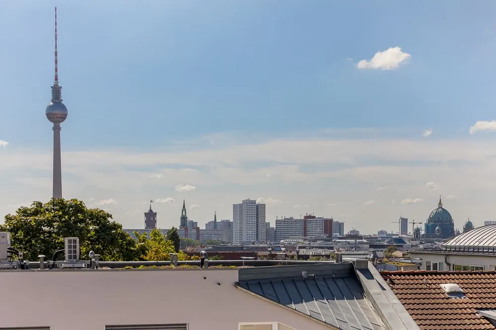 Blick über Hausdächer auf Berliner Fernsehturm und Stadtsilhouette bei blauem Himmel.