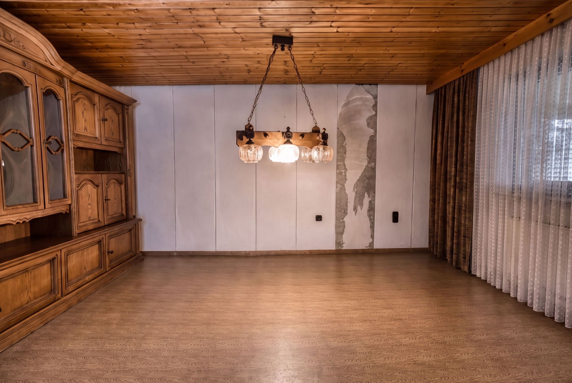 Leeres Zimmer mit Holzdecke, Kronleuchter, Holzschrank und beschädigter Wand.