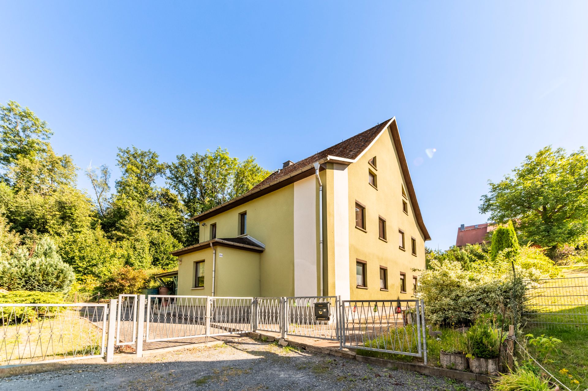 Gelbes Wohnhaus mit Zaun und Garten, umgeben von Bäumen, bei sonnigem Wetter.