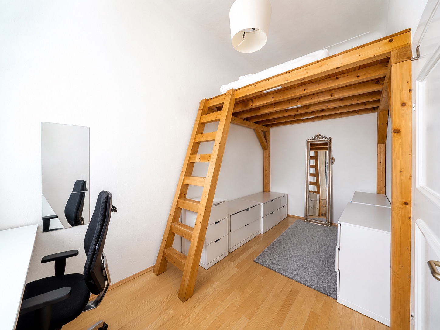 Helles Zimmer mit Hochbett aus Holz, Schreibtisch, Stuhl, Kommoden und einem Spiegel.